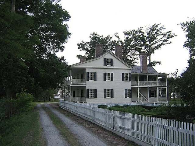 Historic main house at Somerset Place in Creswell, NC, surrounded by a white picket fence, gravel driveway, and mature cypress trees along the waterfront